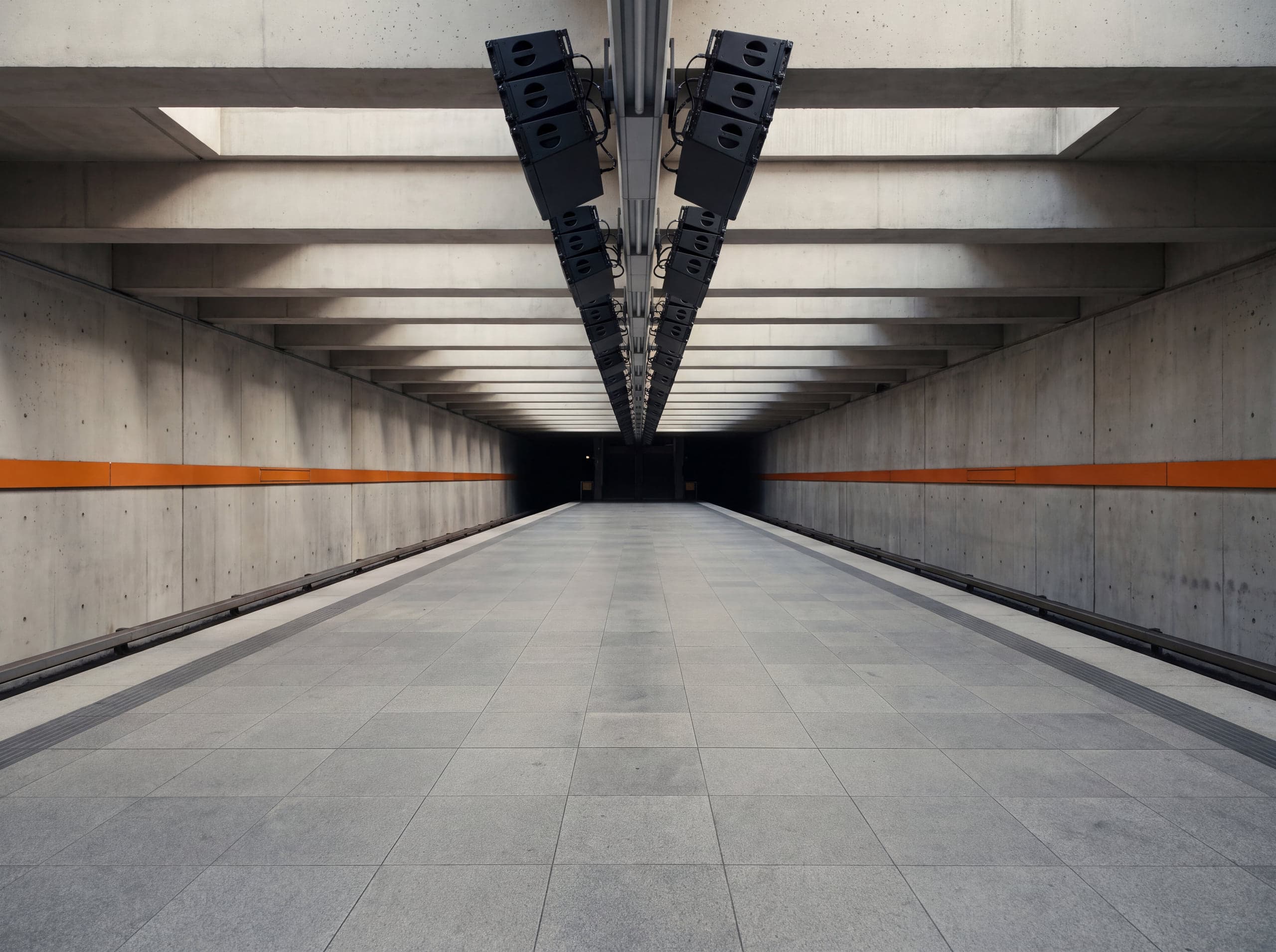 Unterirdischer Bahnsteig mit Lautsprecherzeile und orangefarbenem Leitstreifen an der Wand